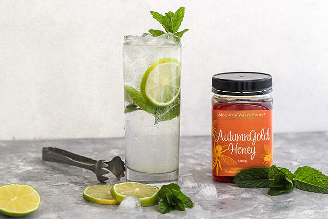 A cool, refreshing mojito in a tall glass with mint leaves and lime slices with a pot of Autumn Gold honey beside it.